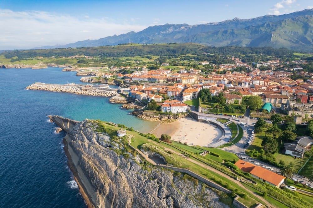 Coastal town of Llanes in Asturias North Spain shown in daytime with traditional buildings along the shoreline and mountains rising in the background.