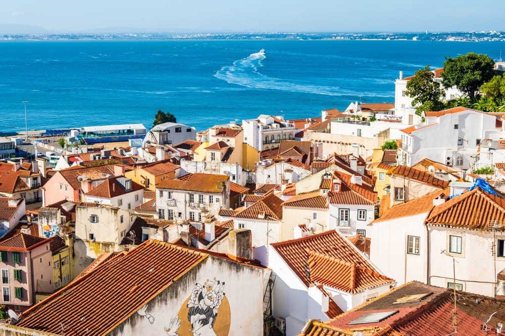 Elevated view of Lisbon’s Alfama district from the Portas do Sol viewpoint showing tightly packed houses with clay orange roofs, narrow winding streets and the cityscape stretching towards the bright sky.